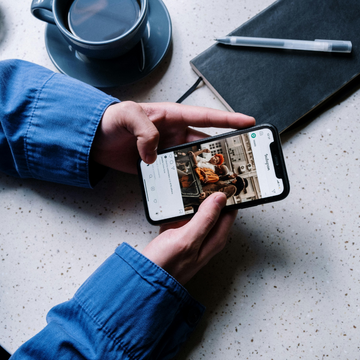 Eine Person hält ein Smartphone auf einem Tisch mit Notizbuch und Kaffeetasse.