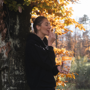 Eine Frau isst Nüsse im herbstlichen Wald, angelehnt an einen Baum.