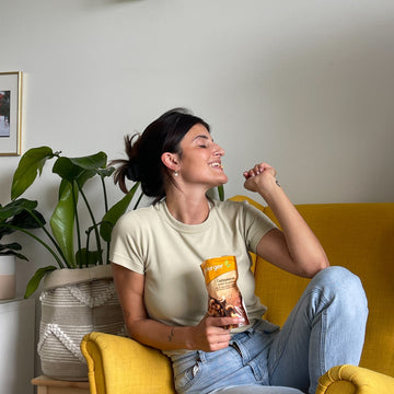 Person isst Cashewkerne aus einer Tüte auf einem gelben Sessel neben einer Pflanze.