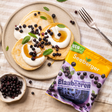 Packung Blaubeeren liegt neben Teller mit Pancakes, die mit Blaubeeren dekoriert wurden
