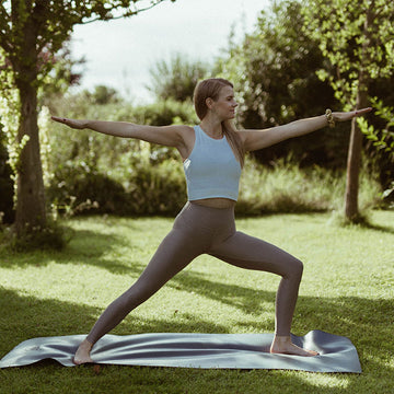Frau macht Yoga-Übung auf Matte, auf einer Wiese, umgeben von Bäumen.