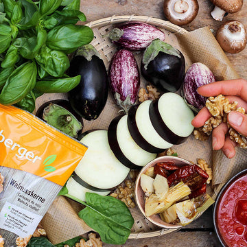 Auberginen, Walnusshälften und andere Zutaten liegen dekorativ auf einem Tisch neben einem Basilikumstrauch.