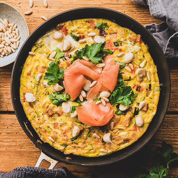 Omelett liegt in Pfanne, dekoriert mit Lachs, Kräutern und Nüssen, auf Holztisch.